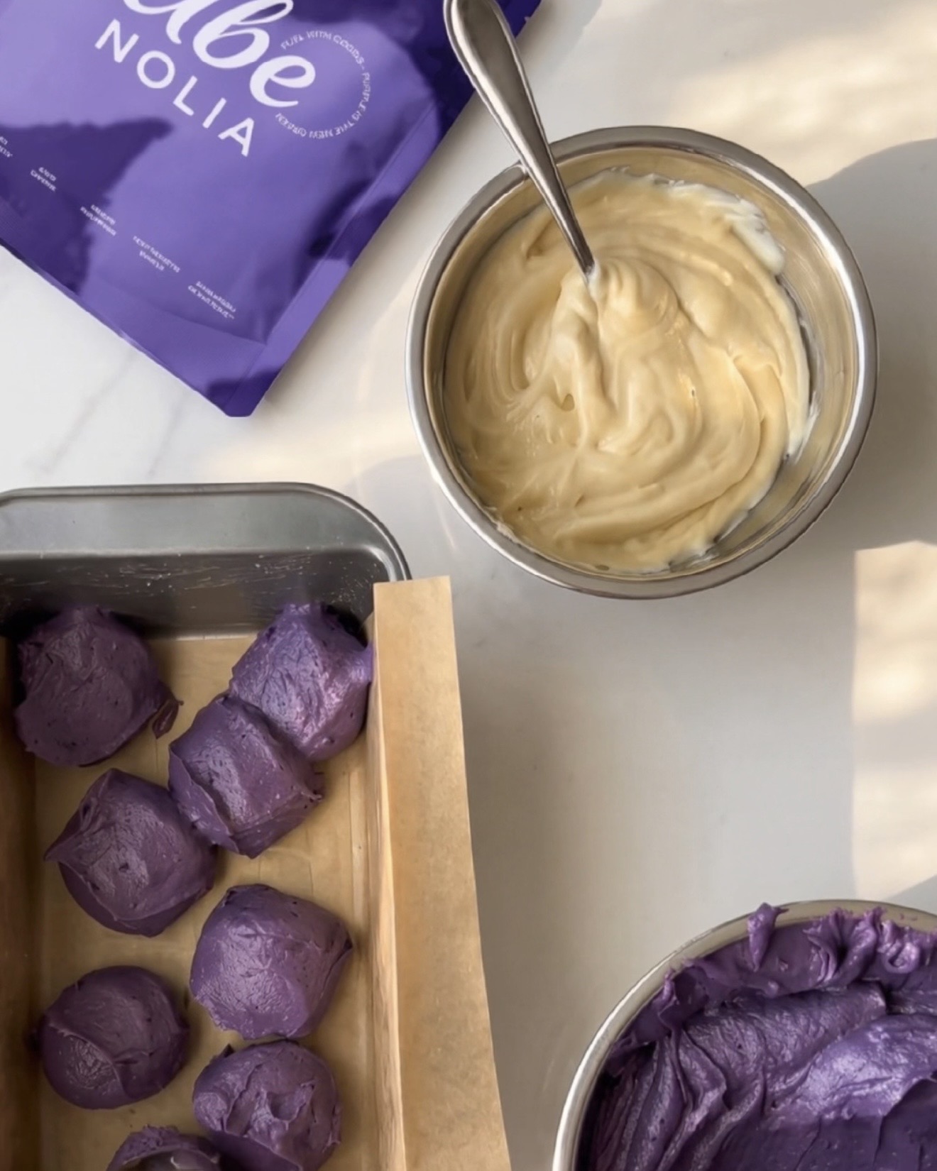 Ube halaya fatto in casa finito, impasto alla vaniglia e cucchiaiate di impasto ube viola in uno stampo da plumcake accanto a una bustina di Ube Nolia polvere di ube