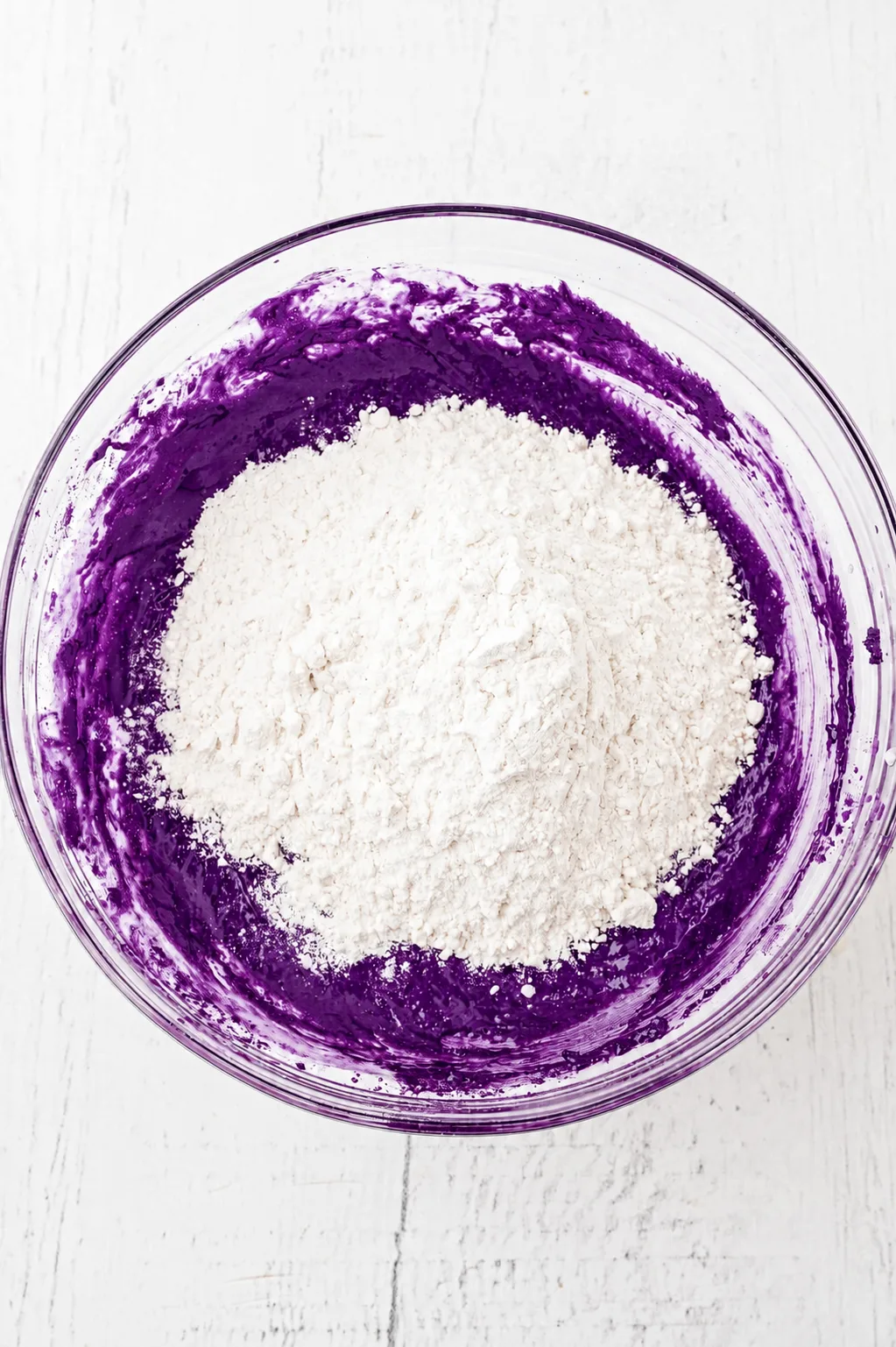 Farina aggiunta sopra l'impasto per brownie ube al cioccolato bianco di colore viola intenso in una ciotola di vetro, pronto per essere incorporato con Ube Nolia powder
