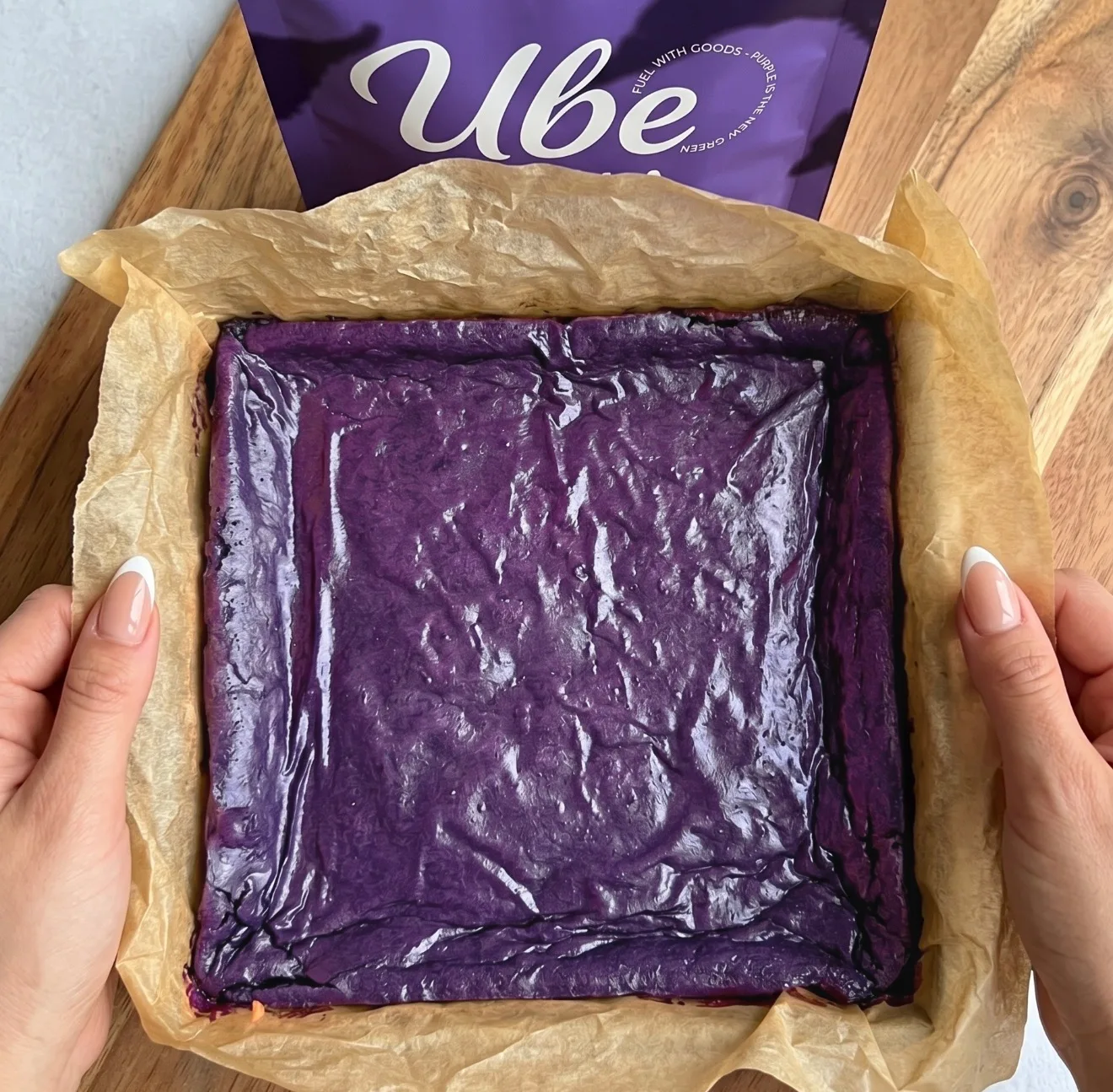 Impasto per brownie ube di colore viola intenso stratificato in una teglia quadrata foderata di carta forno accanto a una bustina di Ube Nolia powder